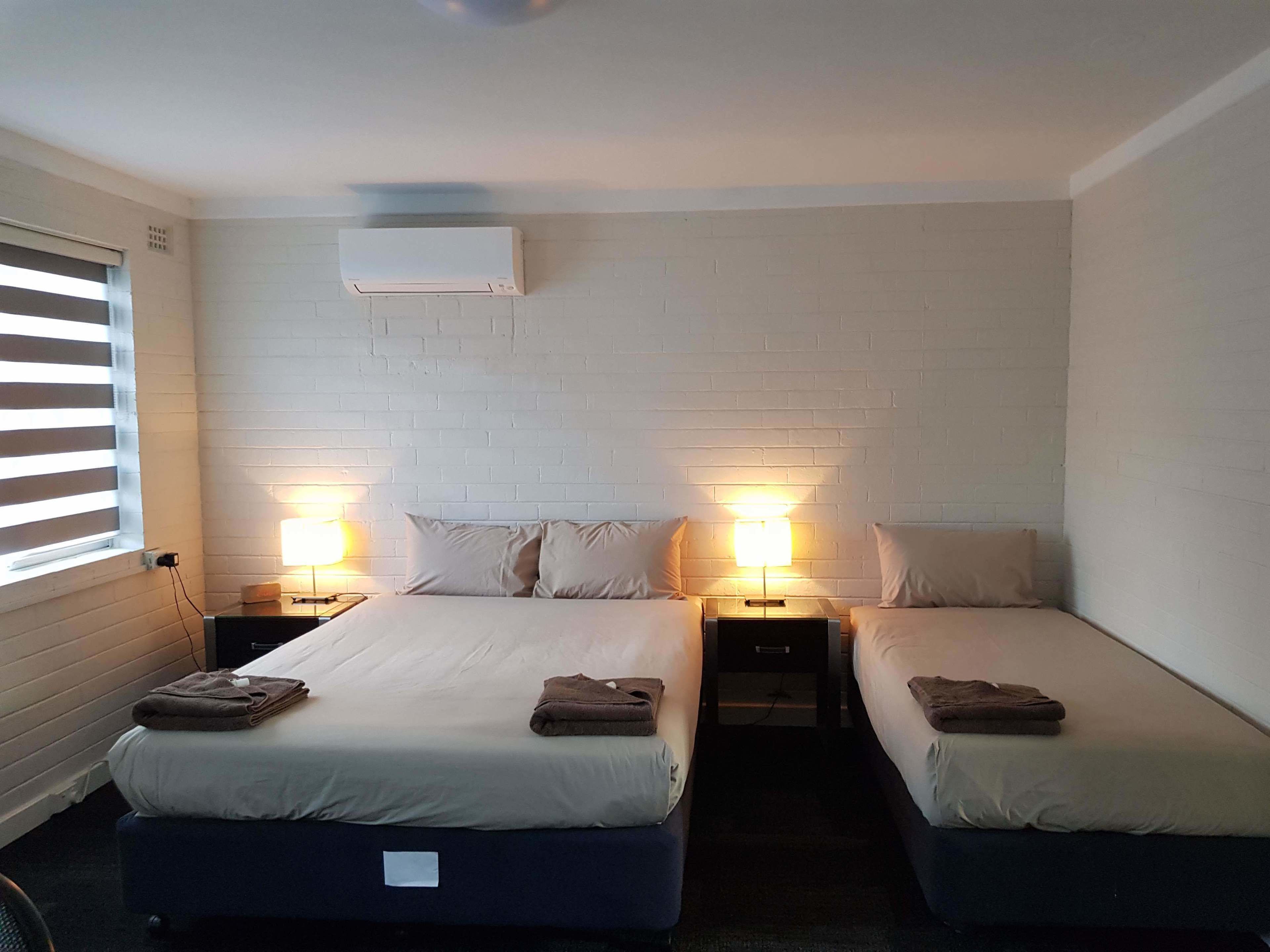 A hotel room with two beds, each with a pillow and a folded towel. There is a wall-mounted air conditioner above the beds and a window with blinds on the left side.