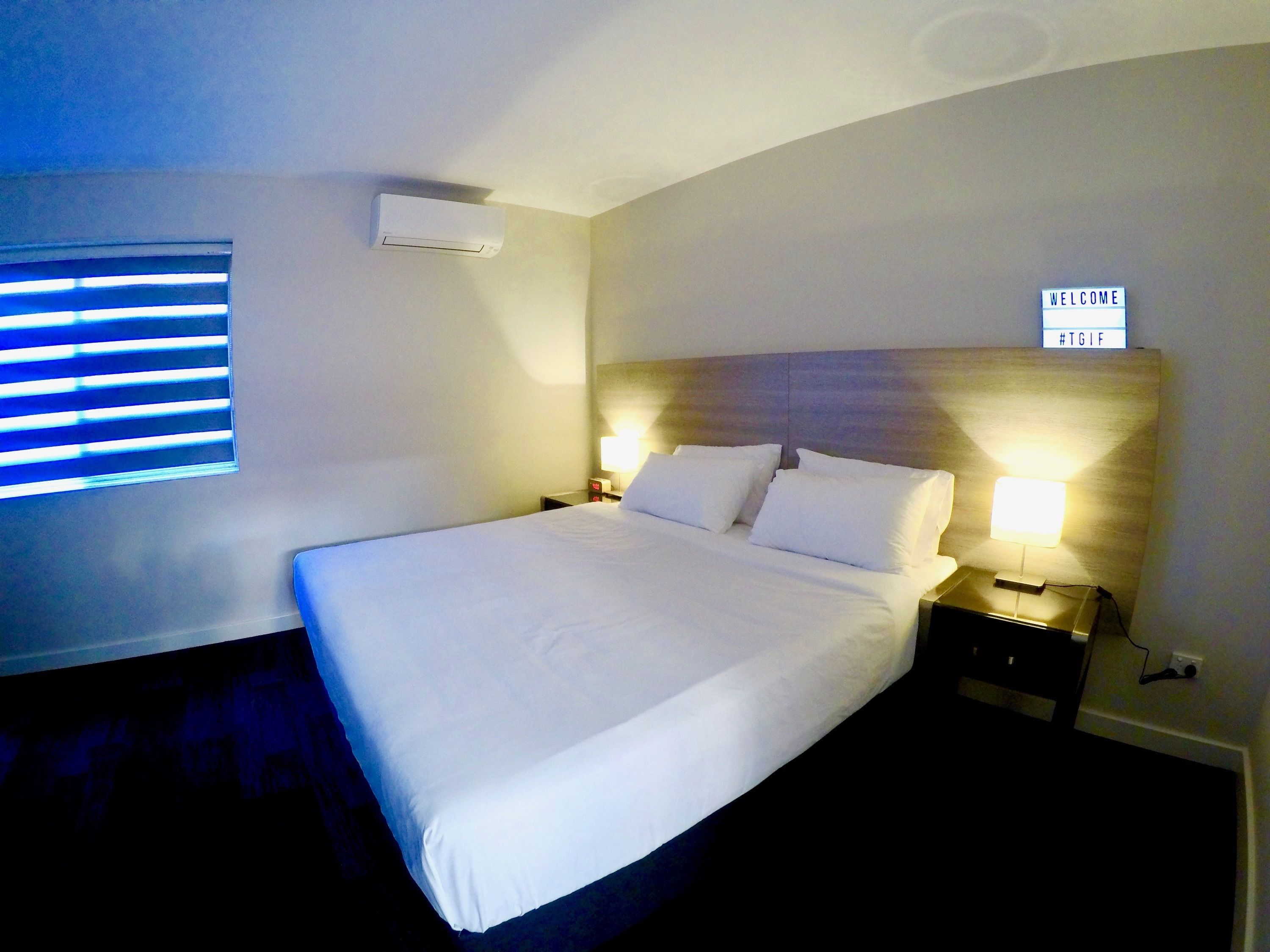 A hotel room with a large bed, two bedside lamps, a wall-mounted air conditioner, and a window with blinds.