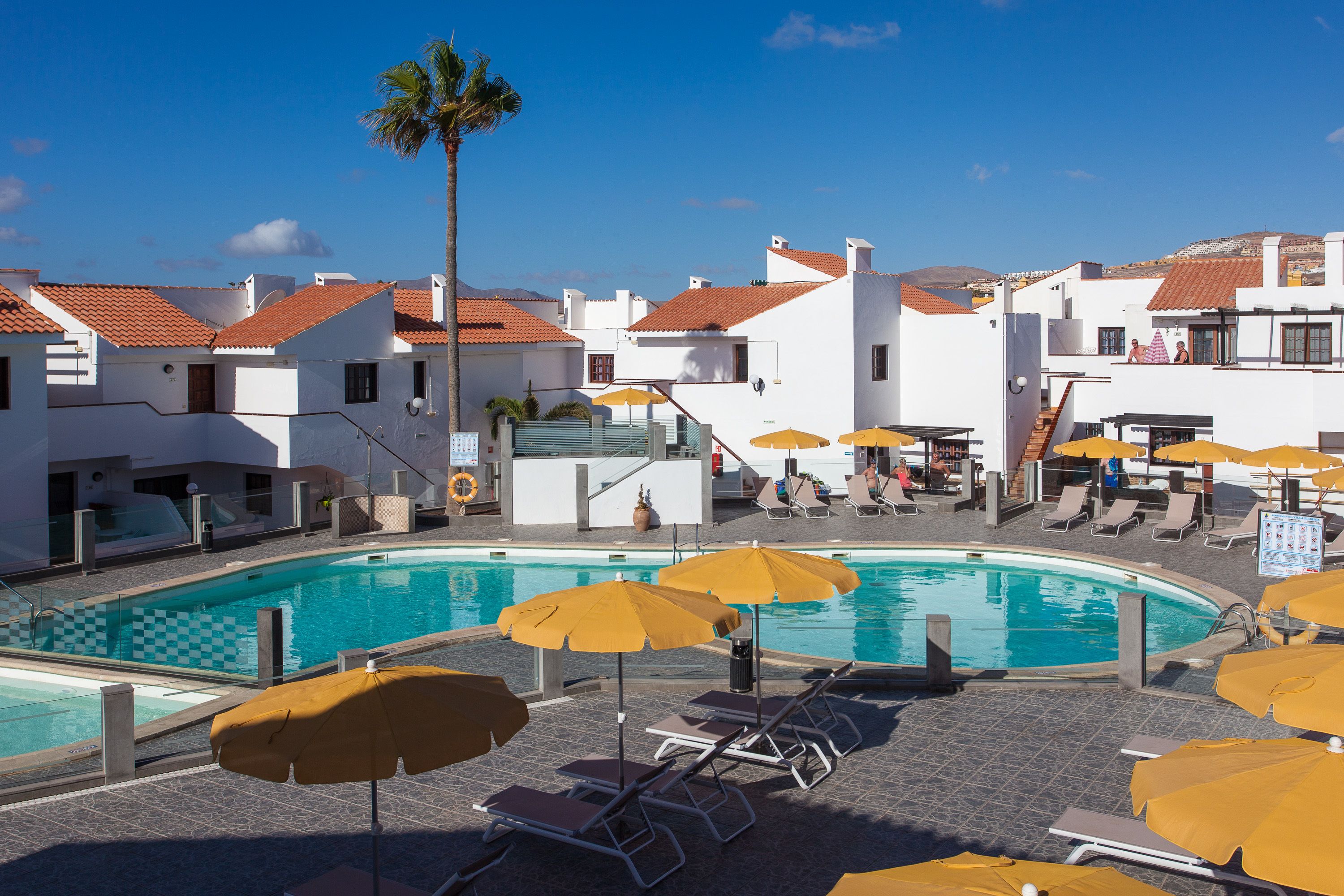 A pool area with sun loungers and umbrellas, surrounded by white buildings with red-tiled roofs.