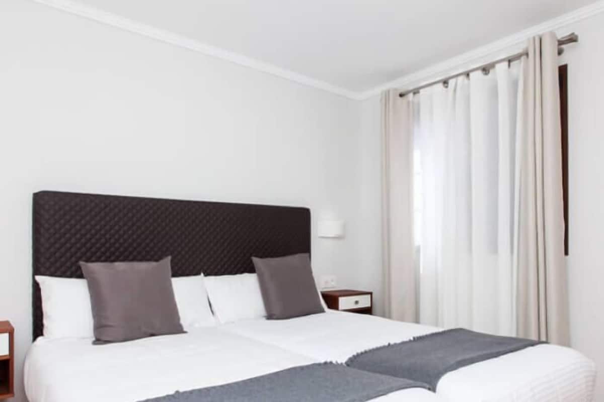 Ein Doppelbett mit gestepptem Kopfteil, zwei Nachttische und ein Fenster mit Vorhängen.