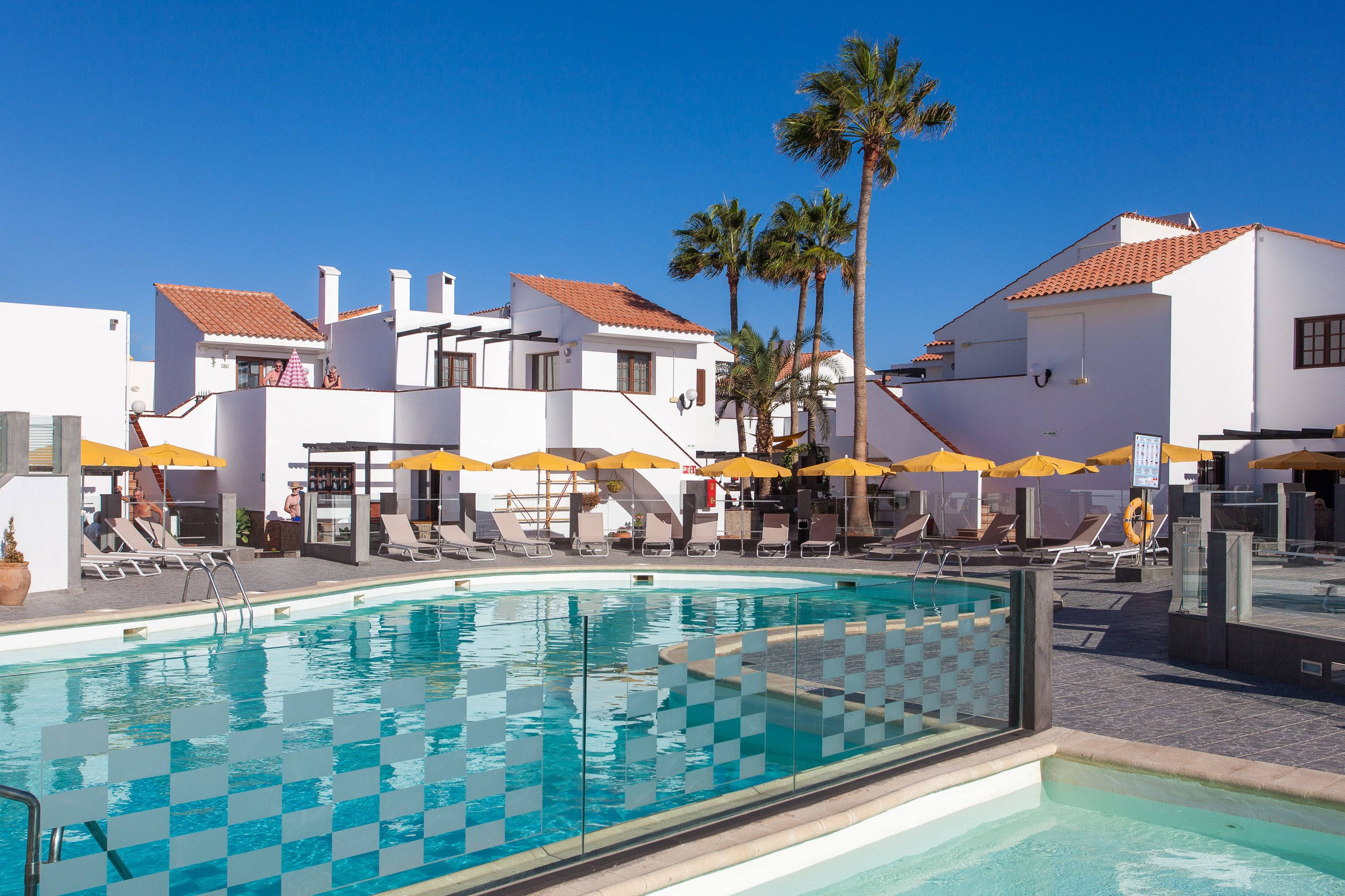 A swimming pool with checkered tiles, surrounded by lounge chairs and yellow umbrellas, with white buildings and palm trees in the background.