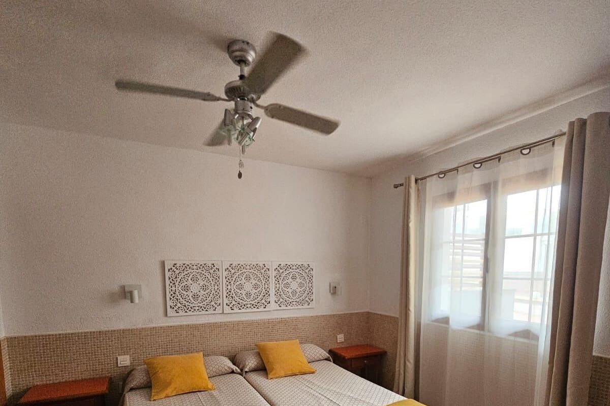 Ein Schlafzimmer mit Bett, Deckenventilator, Nachttisch, einer Wandverkleidung und einem Fenster mit Vorhängen.