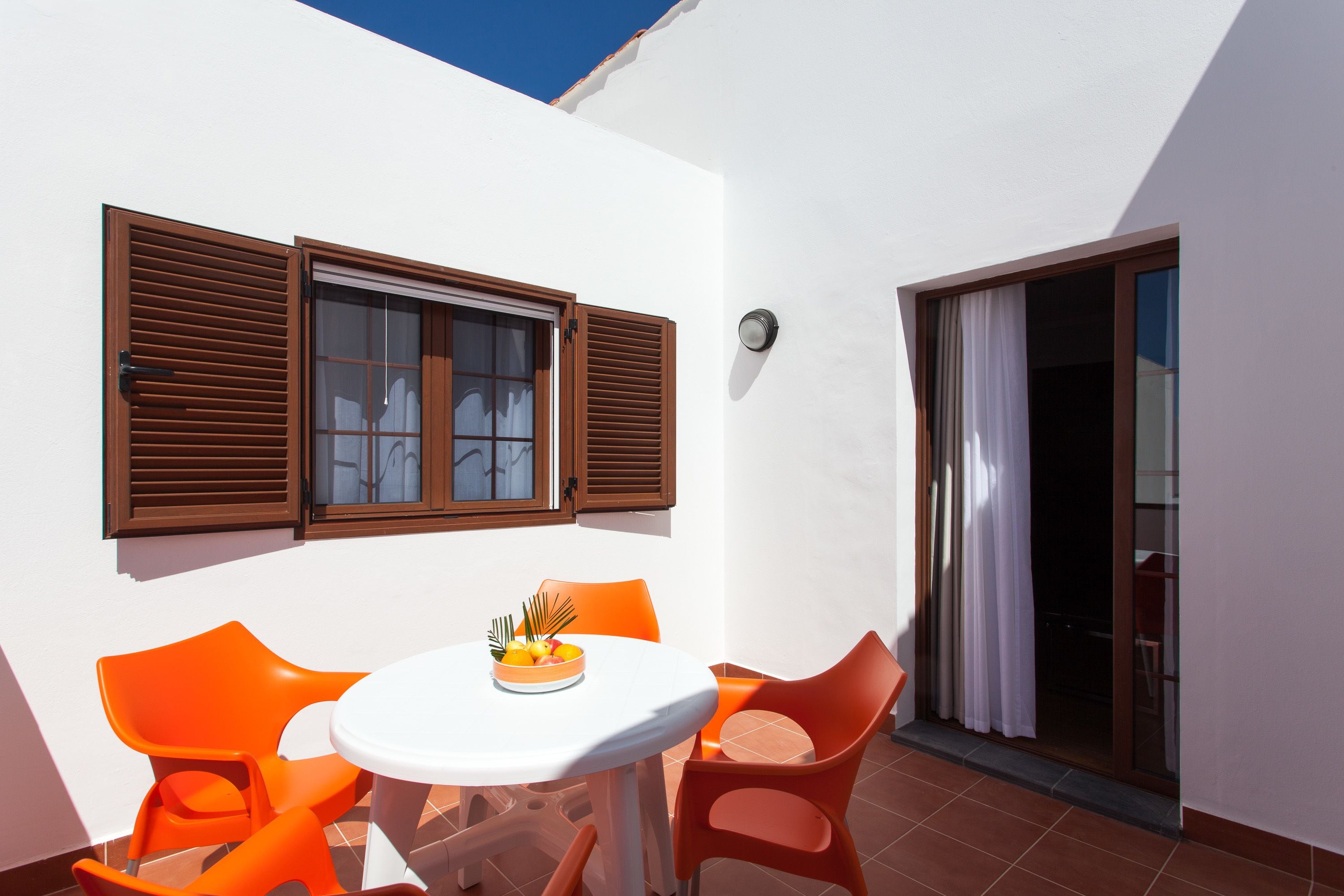 Une petite terrasse avec une table ronde et des chaises orange, une fenêtre avec des volets en bois et une porte-fenêtre coulissante.