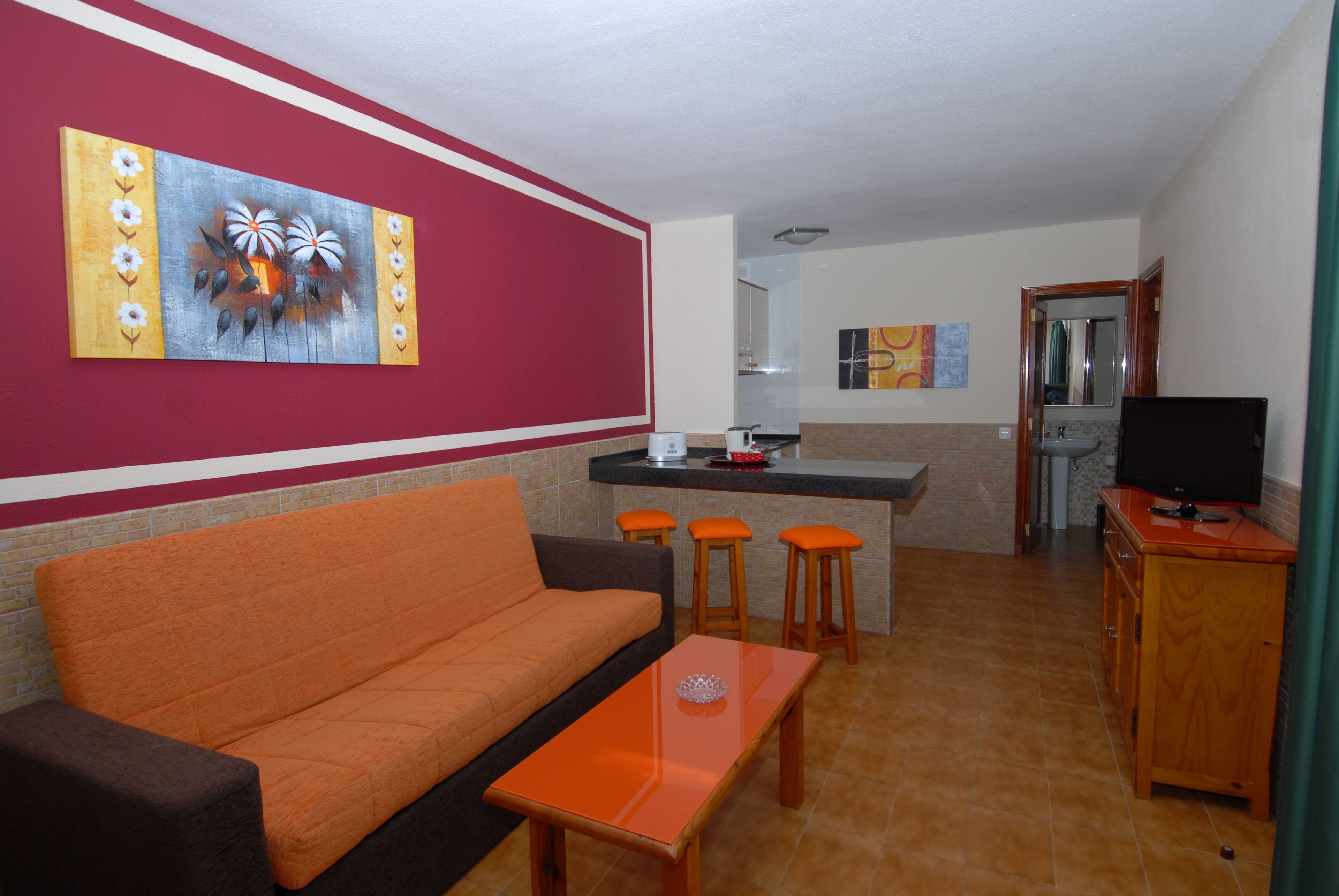 A living room with a sofa, a coffee table, and a TV stand. There is a kitchen area with a counter and stools.