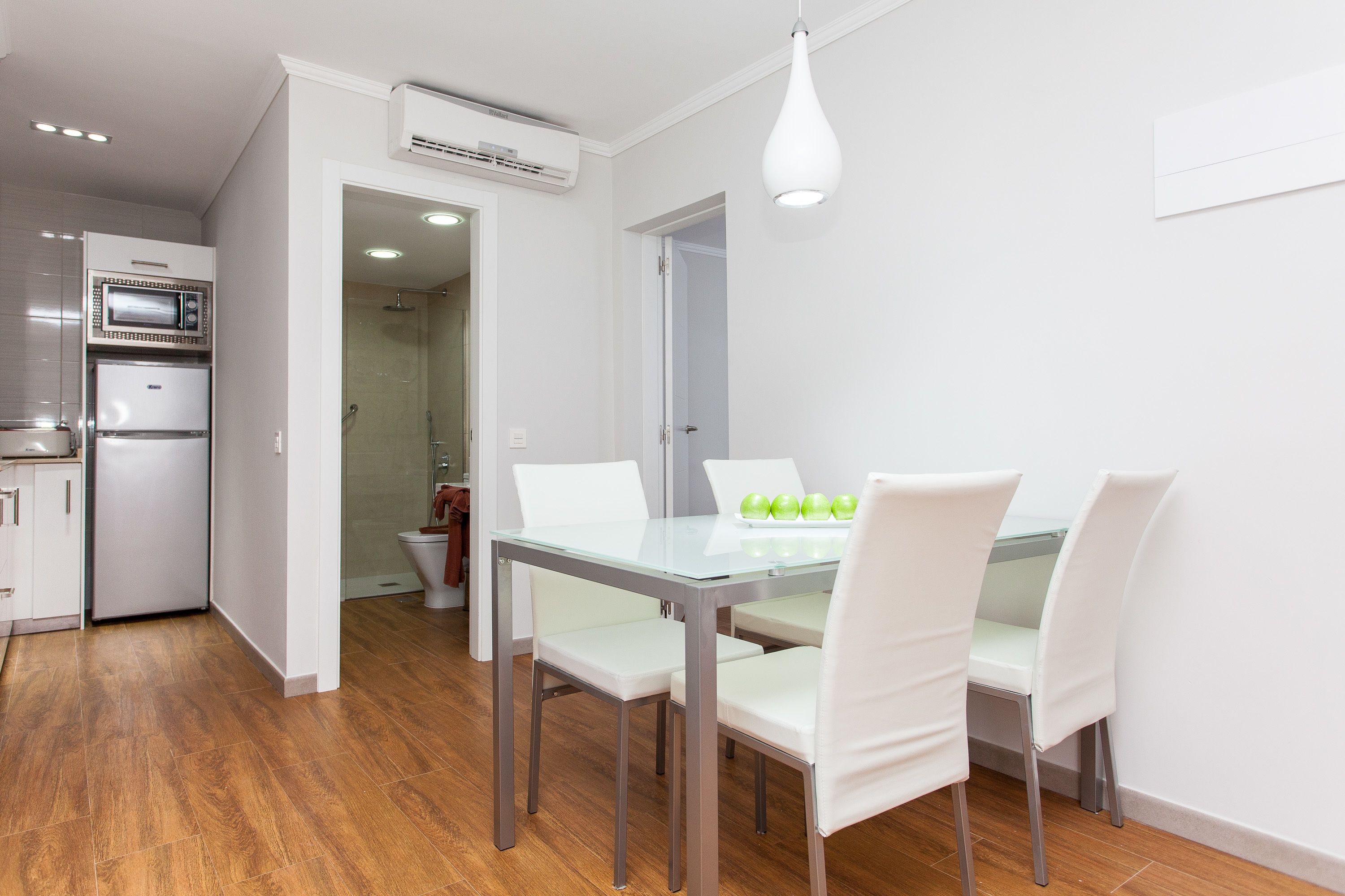 A modern kitchen with a built-in microwave and refrigerator, a dining area with a glass table and white chairs, and a visible shower area in the background.