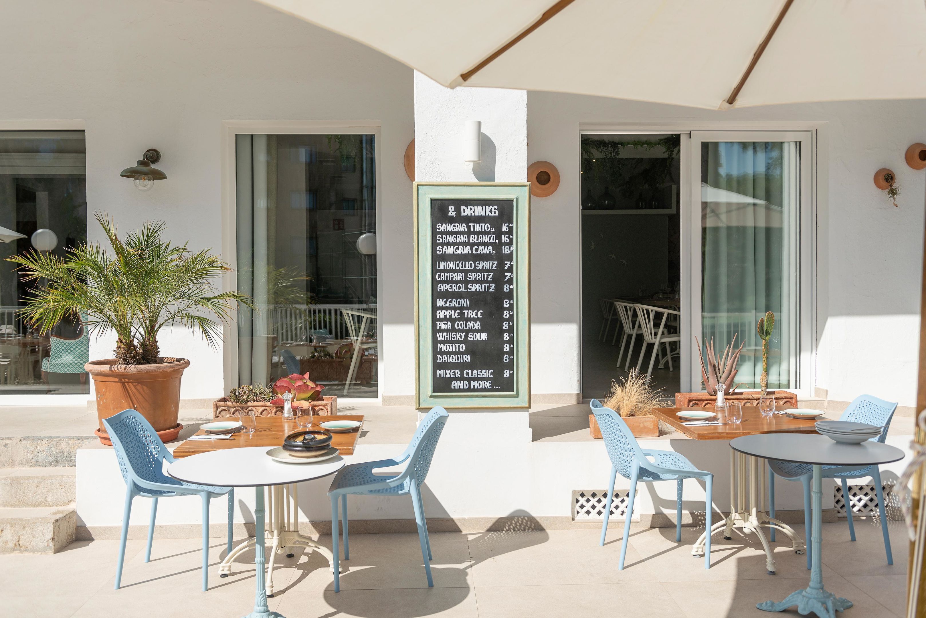 An outdoor seating area with blue chairs and tables, a menu board listing drinks, and a potted plant.