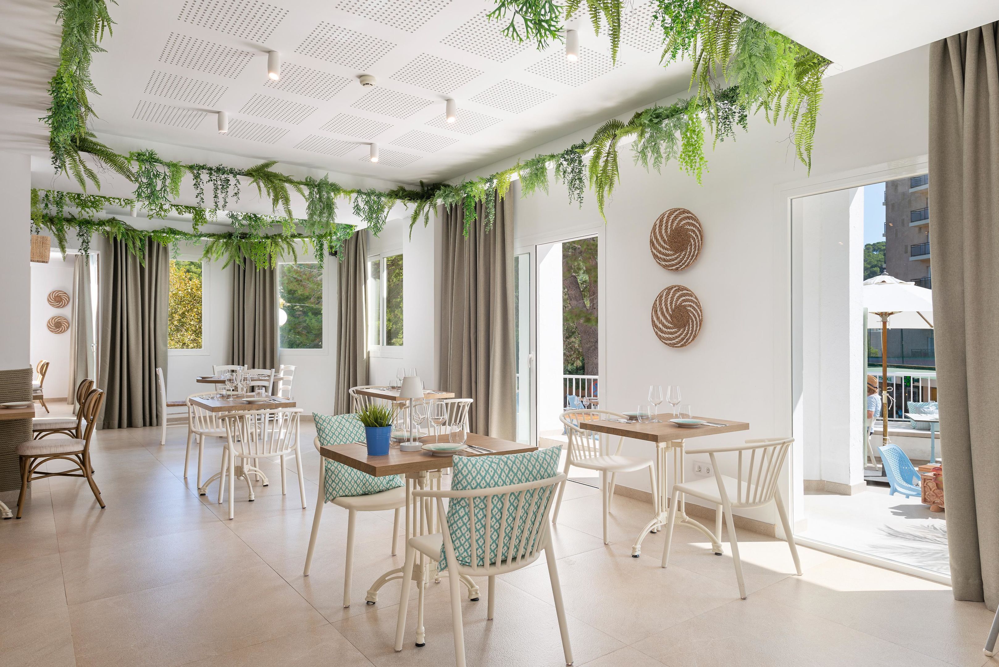 A spacious dining area with white chairs and tables, large windows with curtains, and a ceiling with green foliage.