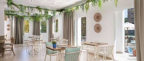 A spacious dining area with white chairs and tables, large windows with curtains, and a ceiling with green foliage.