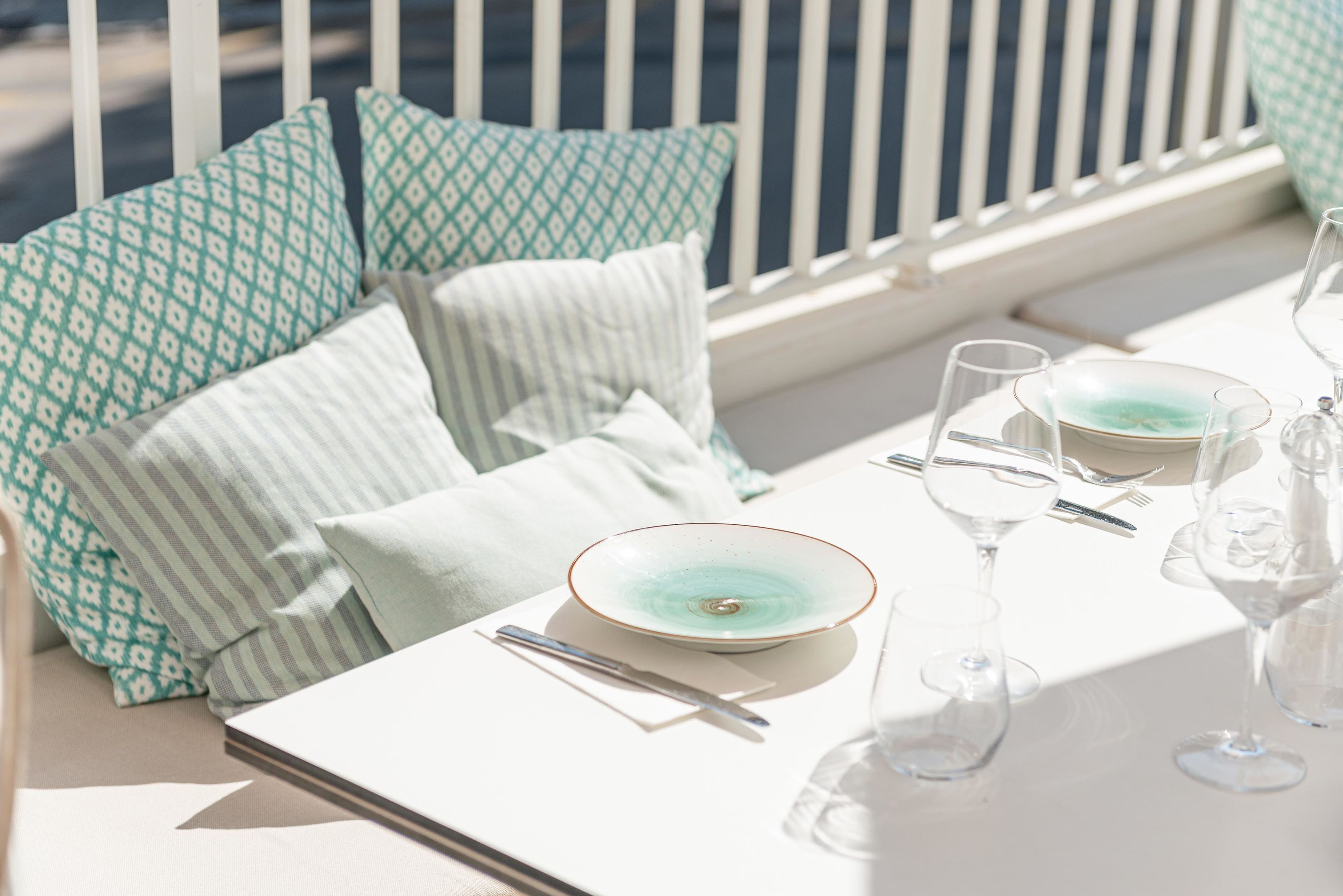A sunlit outdoor seating area with a white table set for two, featuring a teal-patterned cushion and a striped pillow.