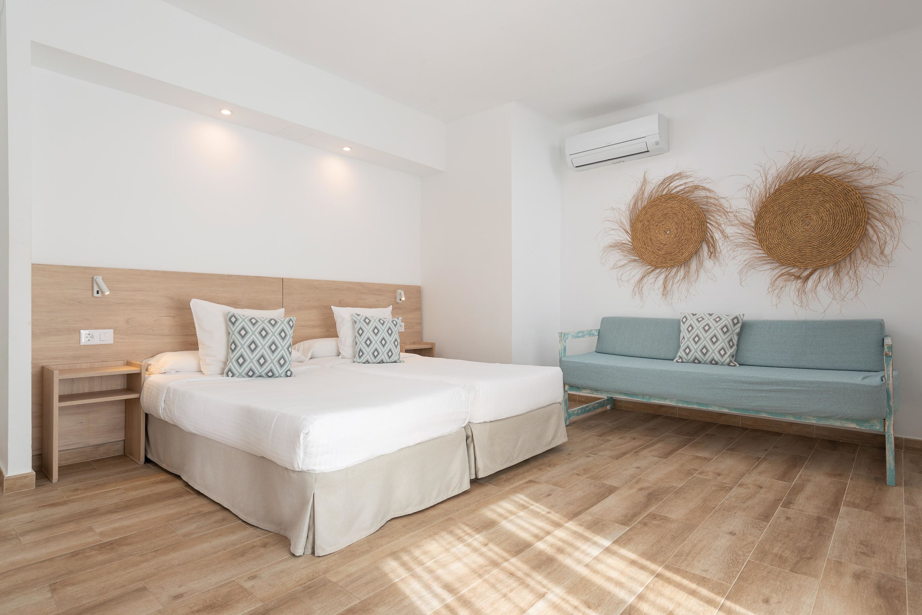 A bedroom with a double bed, bedside tables, a sofa, and two wall-mounted air conditioning units.