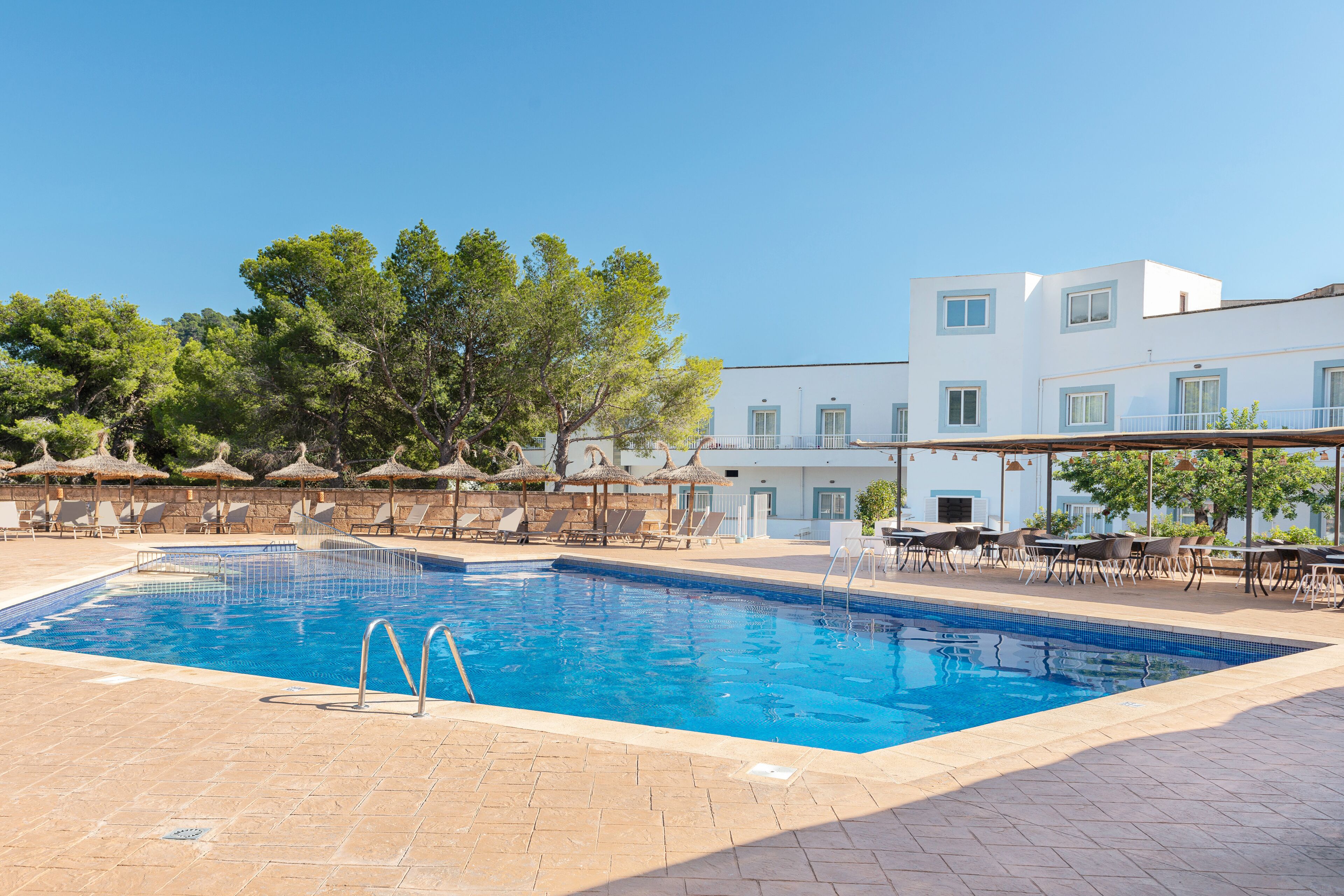 Une piscine entourée de chaises longues et de parasols, le tout bordé d’un bâtiment à la façade blanche et aux fenêtres bleues.