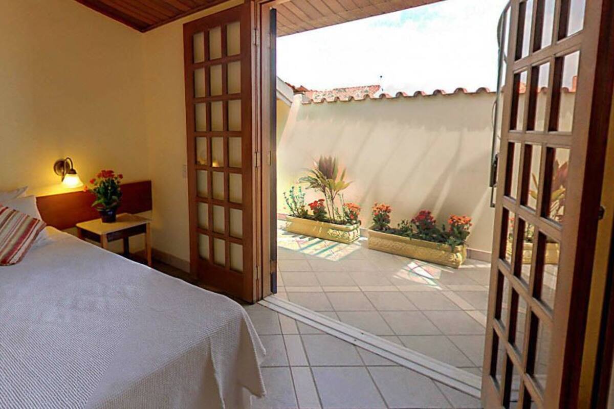 A hotel room with a bed, bedside table, lamp, and an open door leading to a balcony with potted plants.