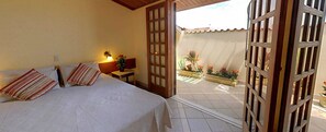 Habitación de hotel con cama, mesita de noche, lámpara y puerta abierta que da a un balcón con plantas en macetas.