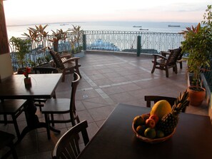 Una terraza con mesas y sillas, un frutero y vistas al mar con barcos.