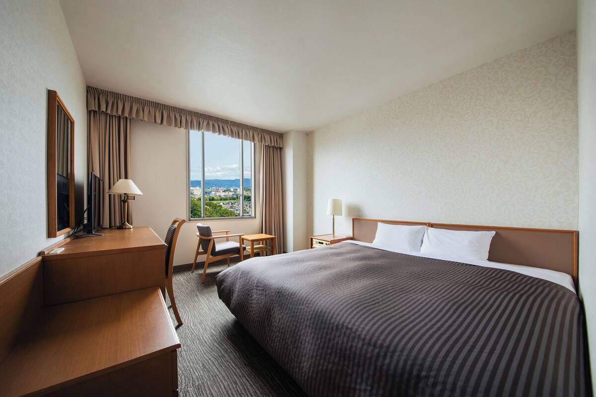 A hotel room with a large bed, a desk, a chair, a lamp, and a view of the city.