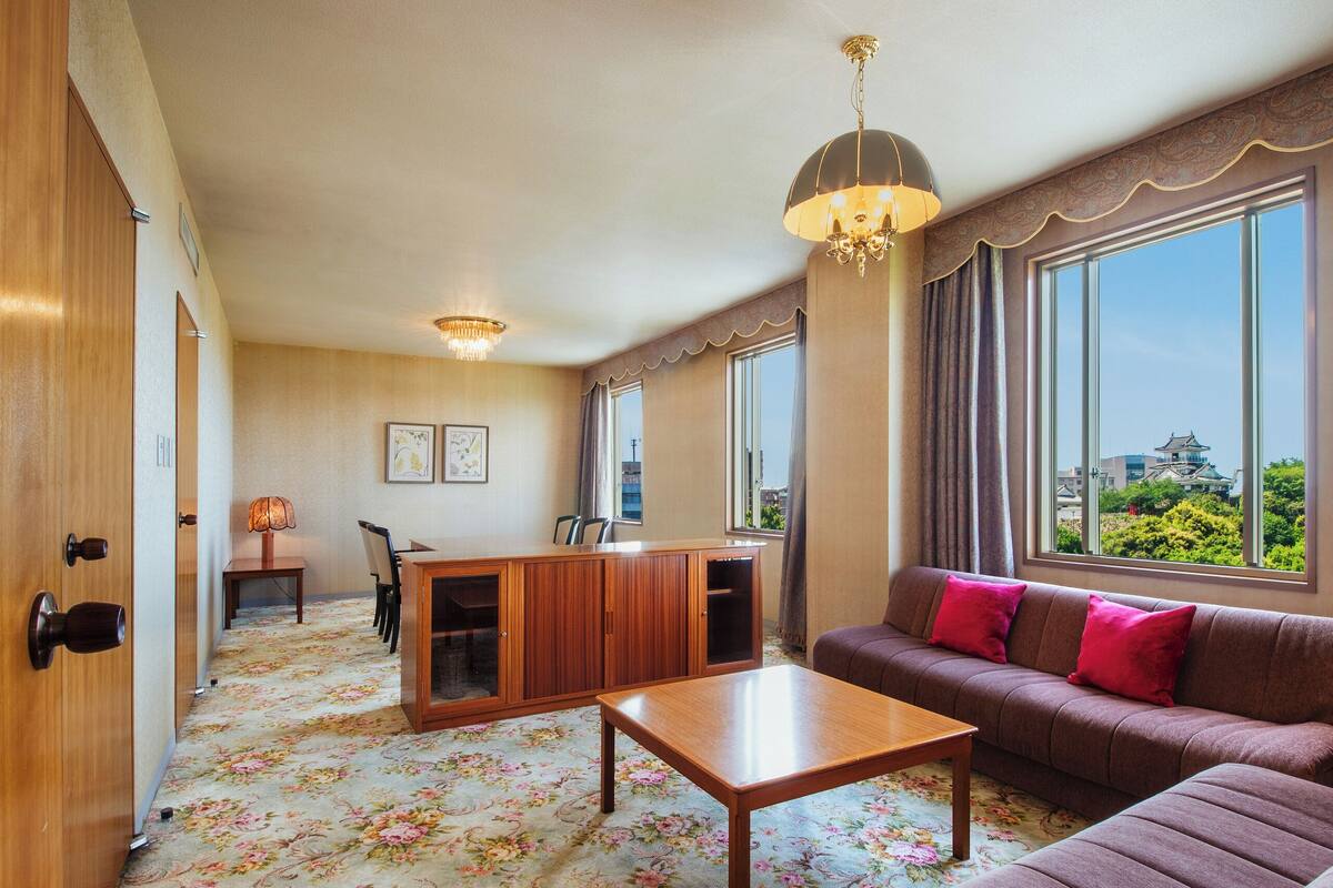 A hotel room with a floral carpet, a sofa with red cushions, a wooden coffee table, a desk with a lamp, and large windows offering a view of greenery and a building.