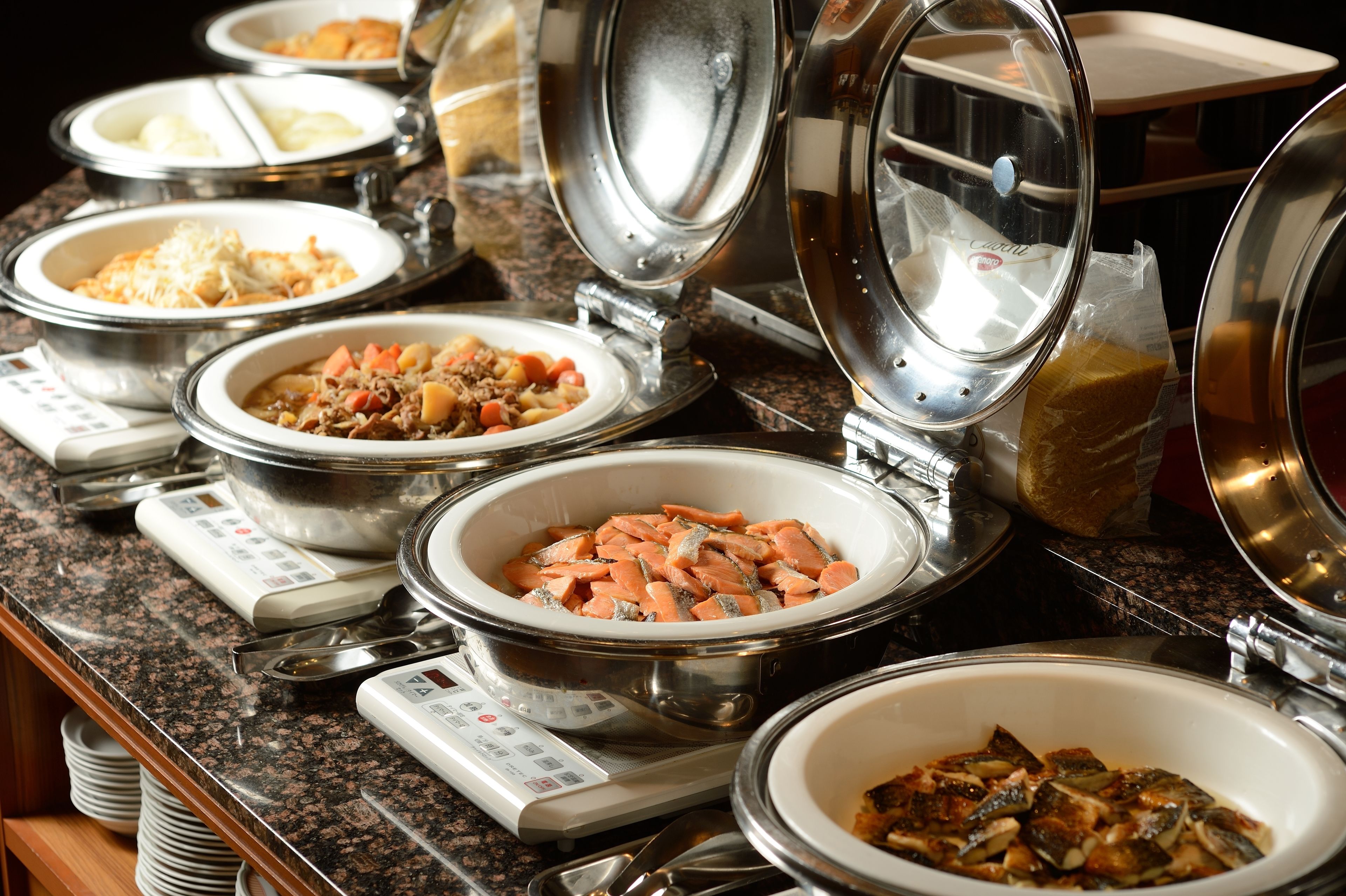 A buffet with various dishes in metal serving dishes, including a bowl of salmon, a bowl of pasta, and a bowl of vegetables.