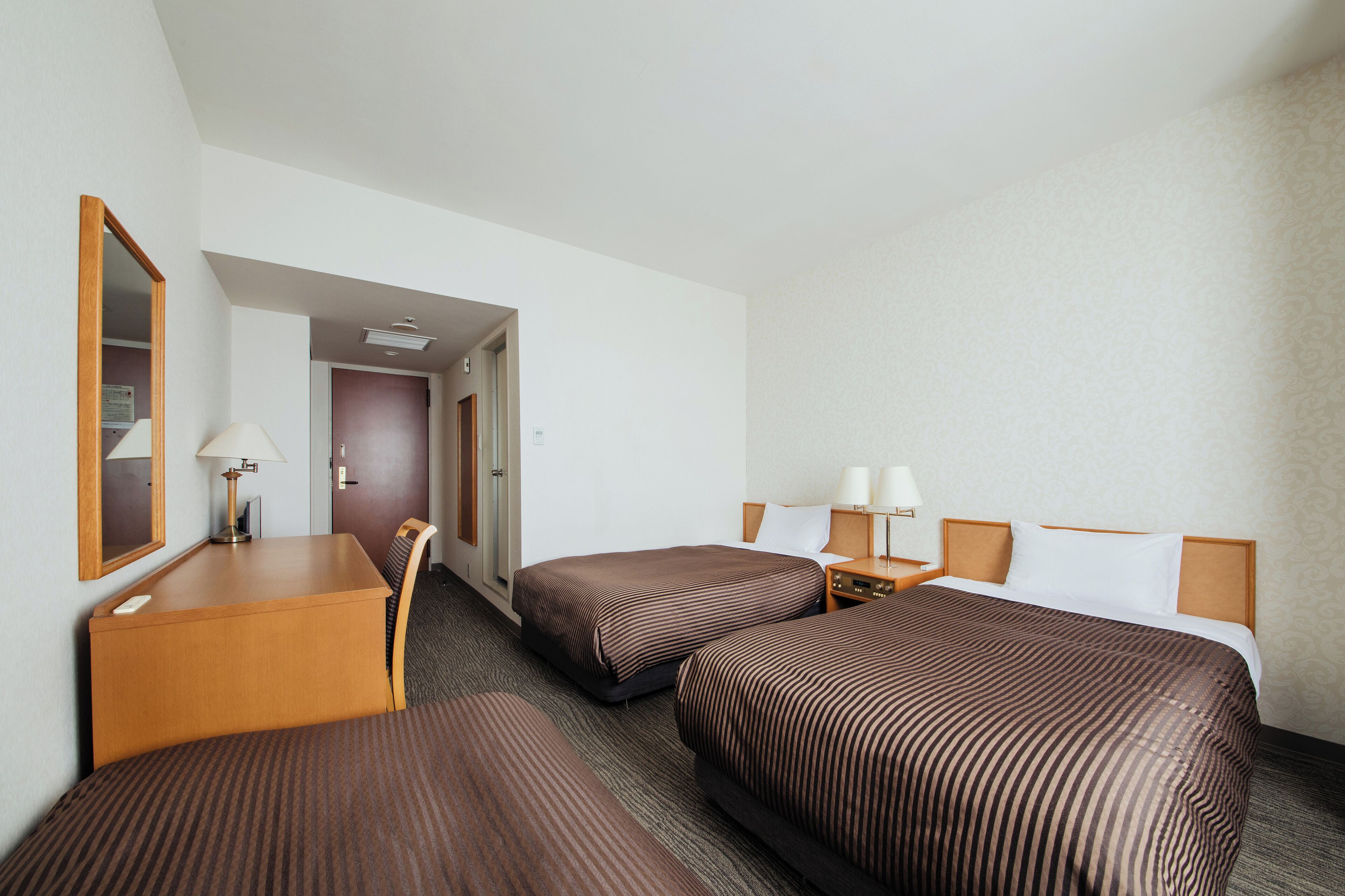Ein Hotelzimmer mit zwei Einzelbetten, einem Schreibtisch mit Stuhl, einem Spiegel und einem Nachttisch mit Lampen.