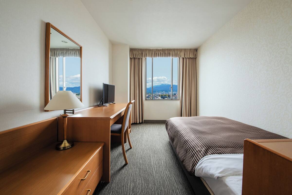 A hotel room with a desk, chair, bed, and a large window offering a city view.
