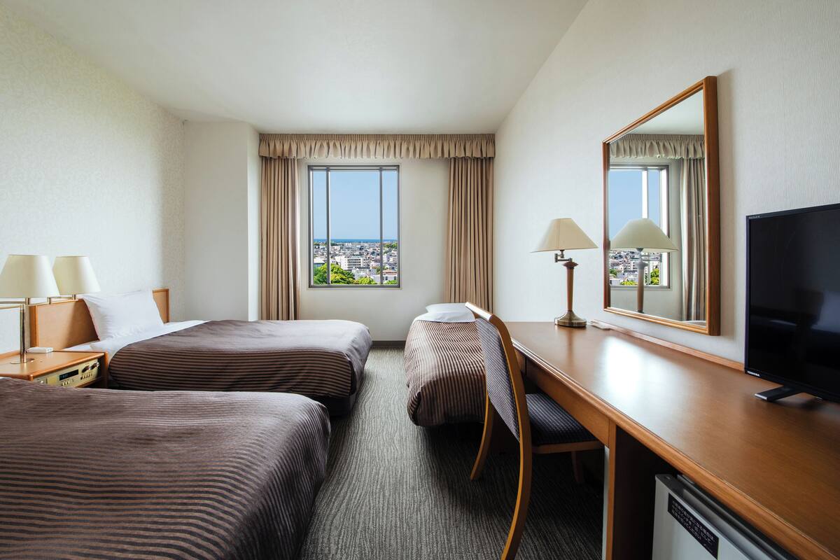 A hotel room with two beds, a desk, a television, and a window with a city view.