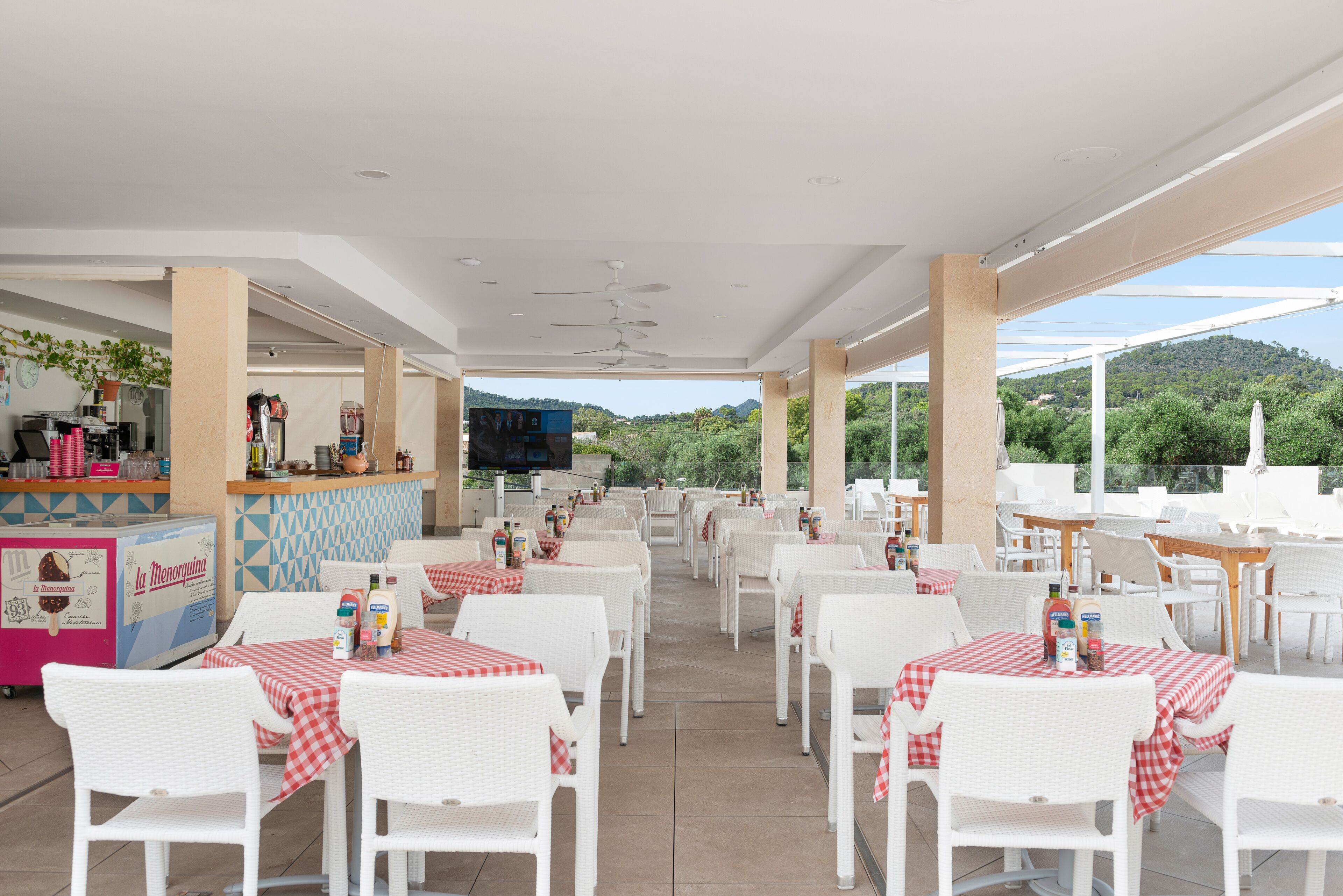 An outdoor dining area with white wicker chairs and tables, a bar area, and a view of greenery and hills.