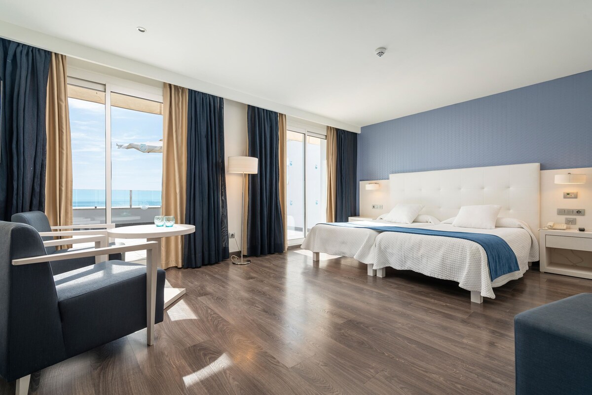 A hotel room with a bed, two bedside tables, a chair, a small table, and a view of the sea through large windows.