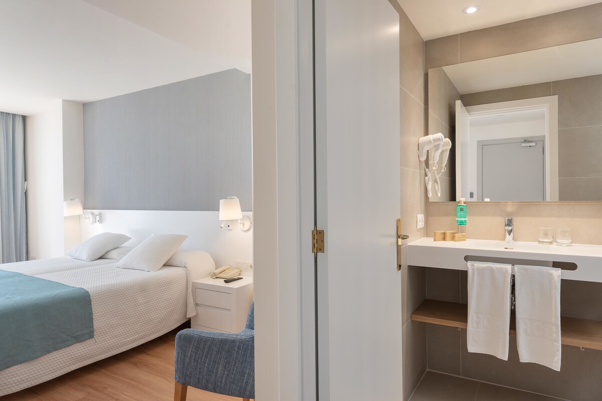 A modern hotel room with a bed, bedside table, lamp, and a bathroom with a sink, mirror, and towel rack.