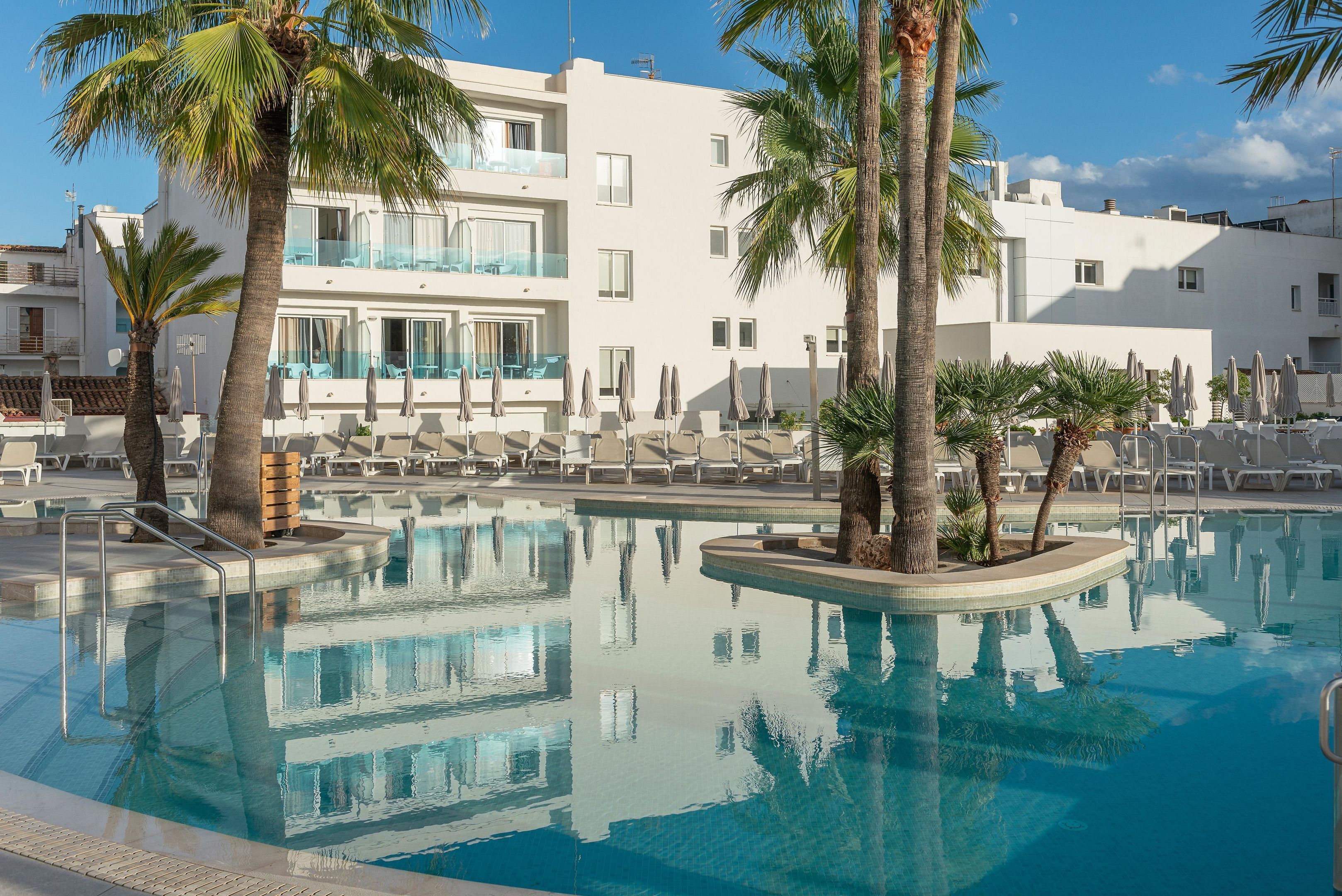 Un área de piscina con sillones de descanso, palmeras y un edificio moderno con balcones.