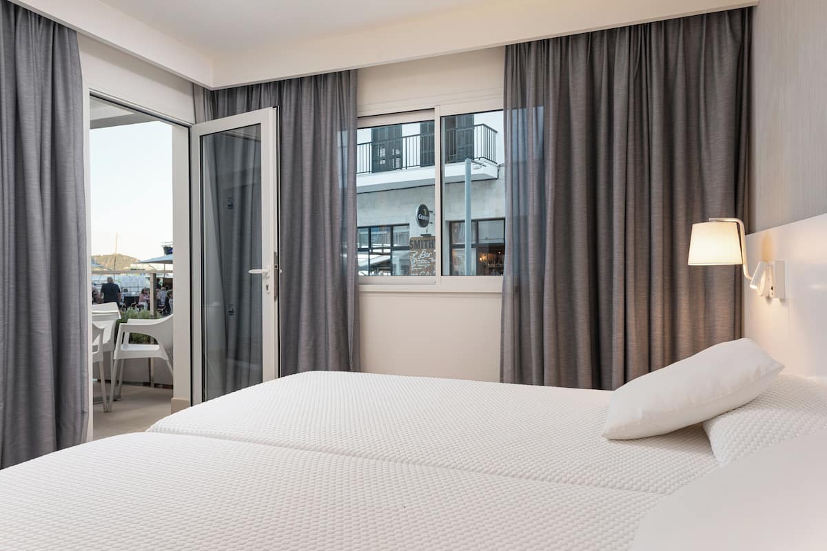 A hotel room with a large bed, grey curtains, a window with a view of the outside, and a small table with chairs.