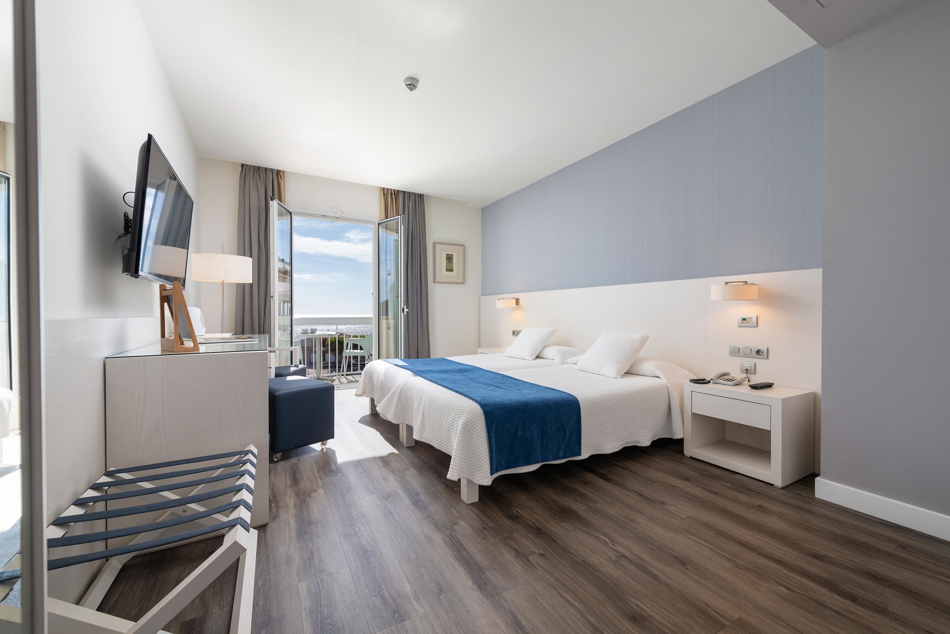A modern hotel room with a large bed, a desk with a lamp, a TV, and a balcony view.