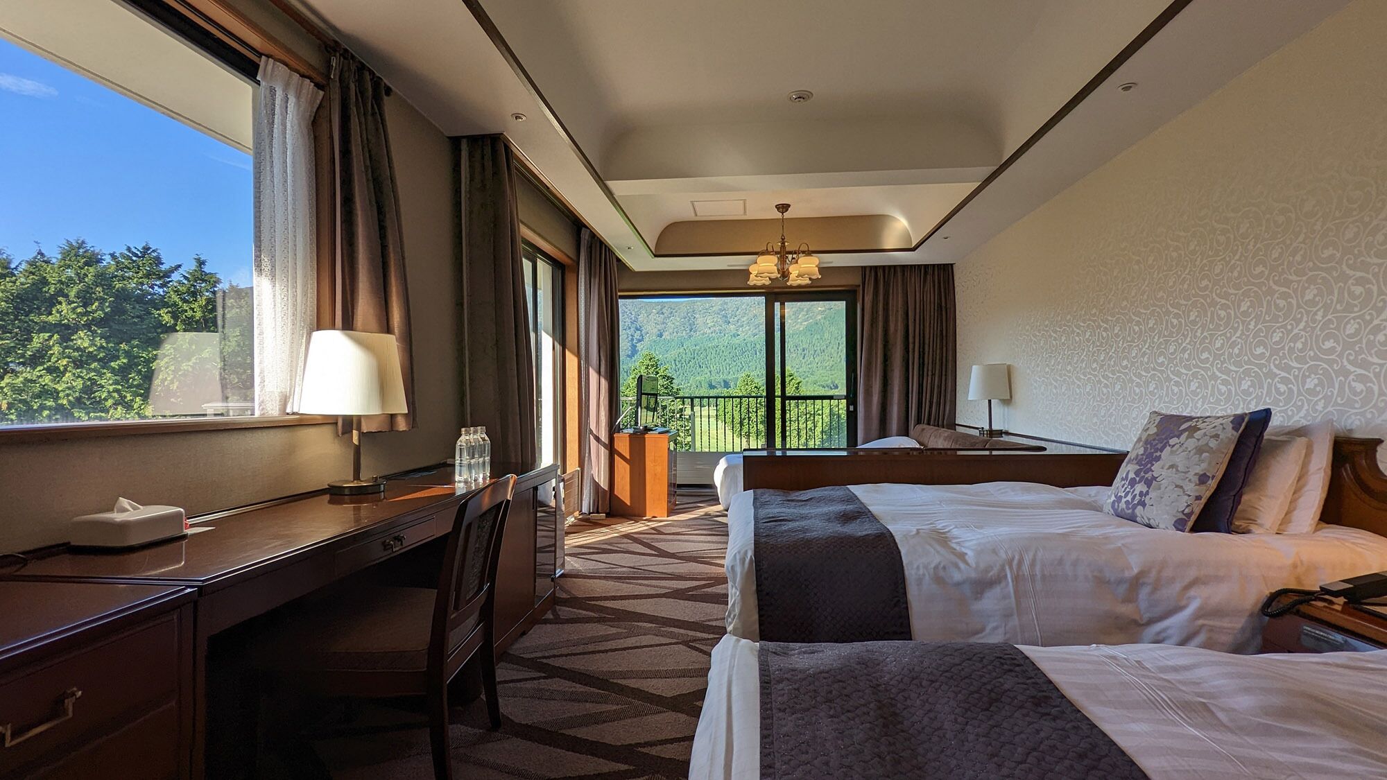 A hotel room with a desk, two beds, a view of greenery, and a balcony.