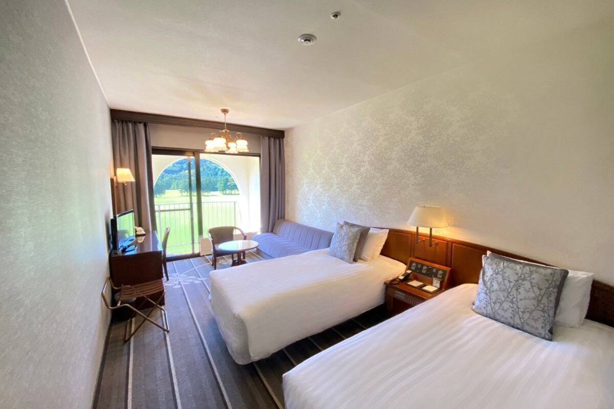 Una camera d'albergo con due letti, una scrivania, una sedia e vista su un paesaggio verde attraverso un'ampia finestra.