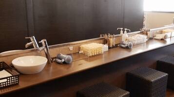 Une salle de bain avec un grand miroir, un lavabo et des articles de toilette posés sur le comptoir. On y trouve des tabourets en osier et un petit panier.