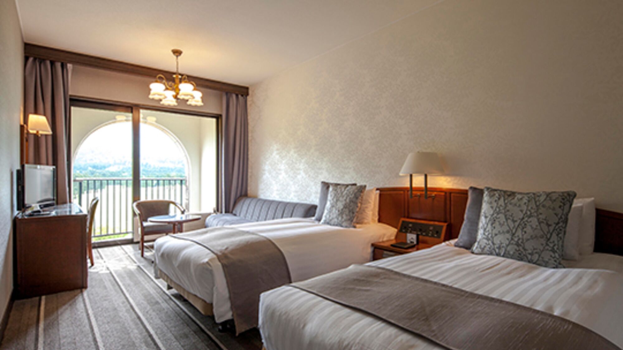 A hotel room with two beds, a desk with a lamp, a chair, and a view of the outdoors.