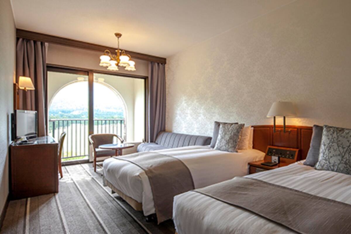 Camera d'albergo con due letti, una scrivania con una lampada, una sedia e vista sull'esterno.