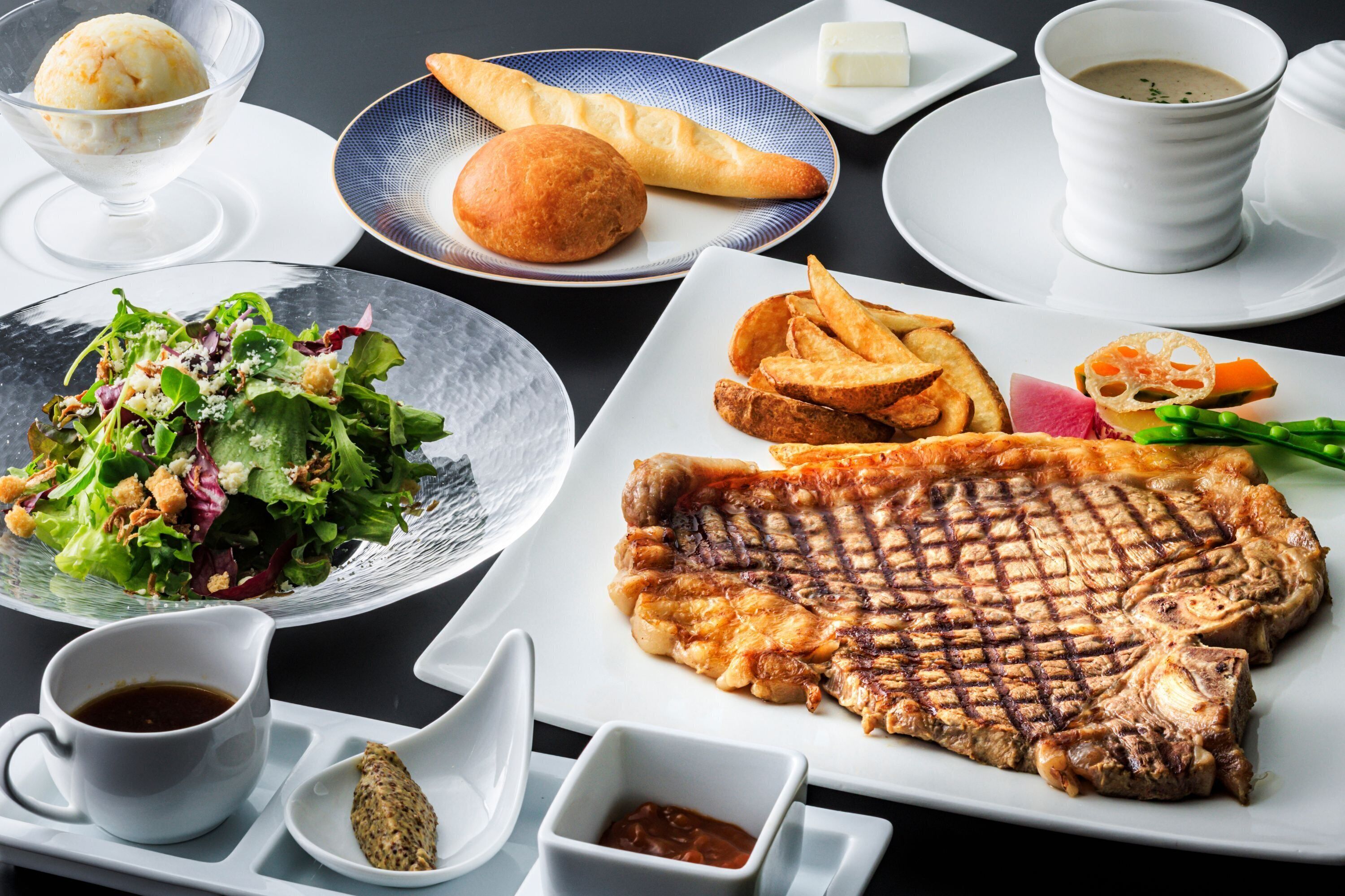 A dining table with a variety of dishes including grilled steak, fries, a salad, and a dessert.
