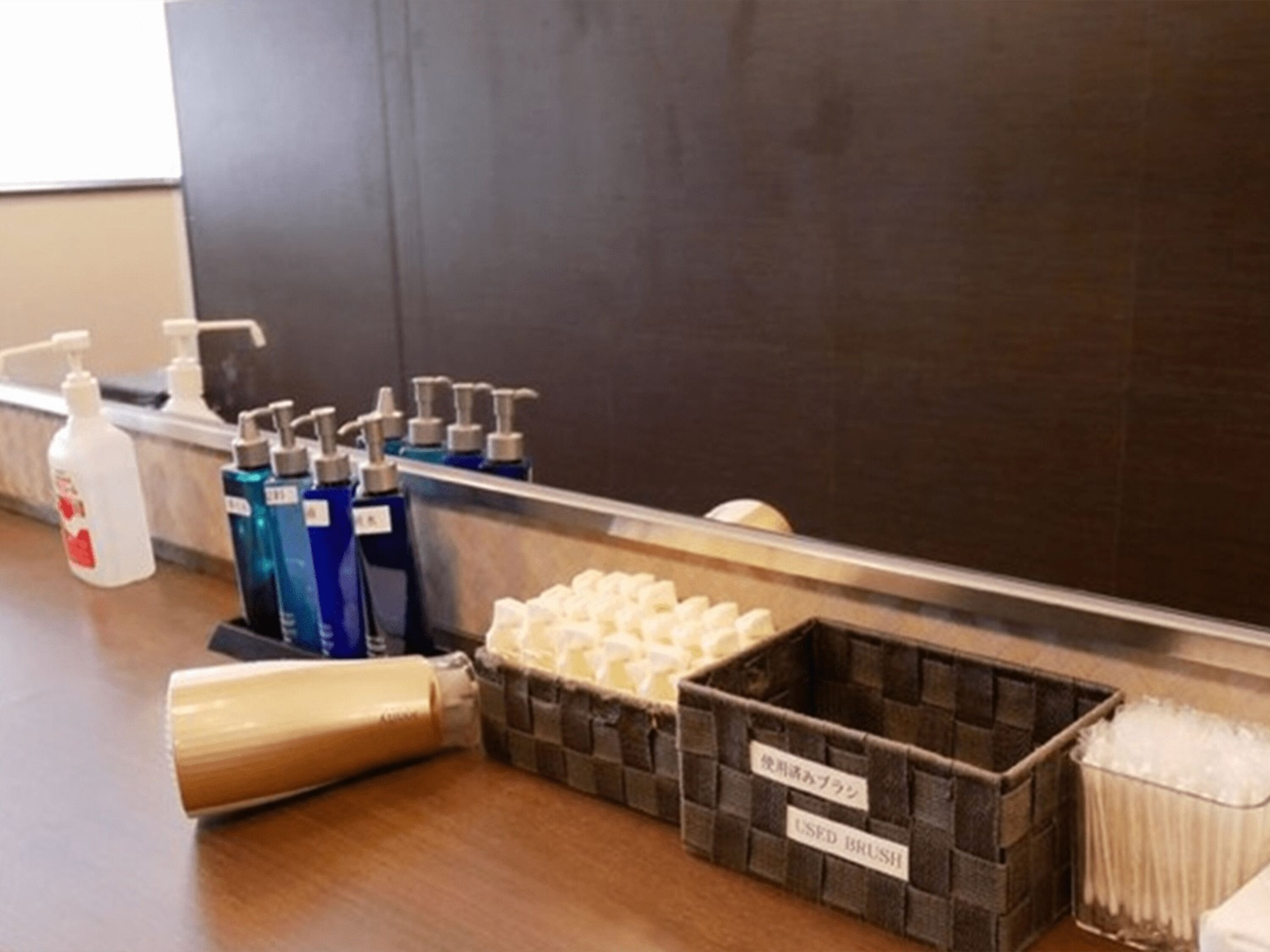 Una encimera de baño con dispensadores de gel hidroalcohólico, un recipiente etiquetado como 'BROCHA USADA' y un cubo de basura.