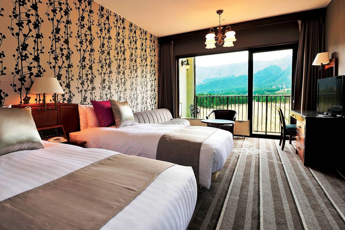 Camera d'albergo con due letti, una scrivania, una sedia e un balcone con vista sulle montagne.