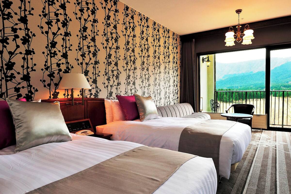 Una camera d'albergo con due letti, una scrivania, una lampada e vista su un balcone con montagne.