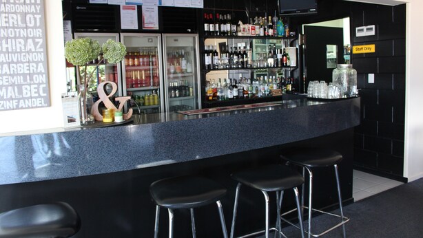 Een bar met een zwart aanrecht, barkrukken en diverse flessen en glazen.
