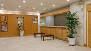 A modern hotel lobby with a wooden reception desk, a large potted plant, and a tiled floor. - Hotel Sunroute Sopra Kobe (Kobe)