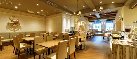 A spacious restaurant with multiple dining tables and chairs, a buffet area with food and beverages, and a view of the outdoors through large windows.