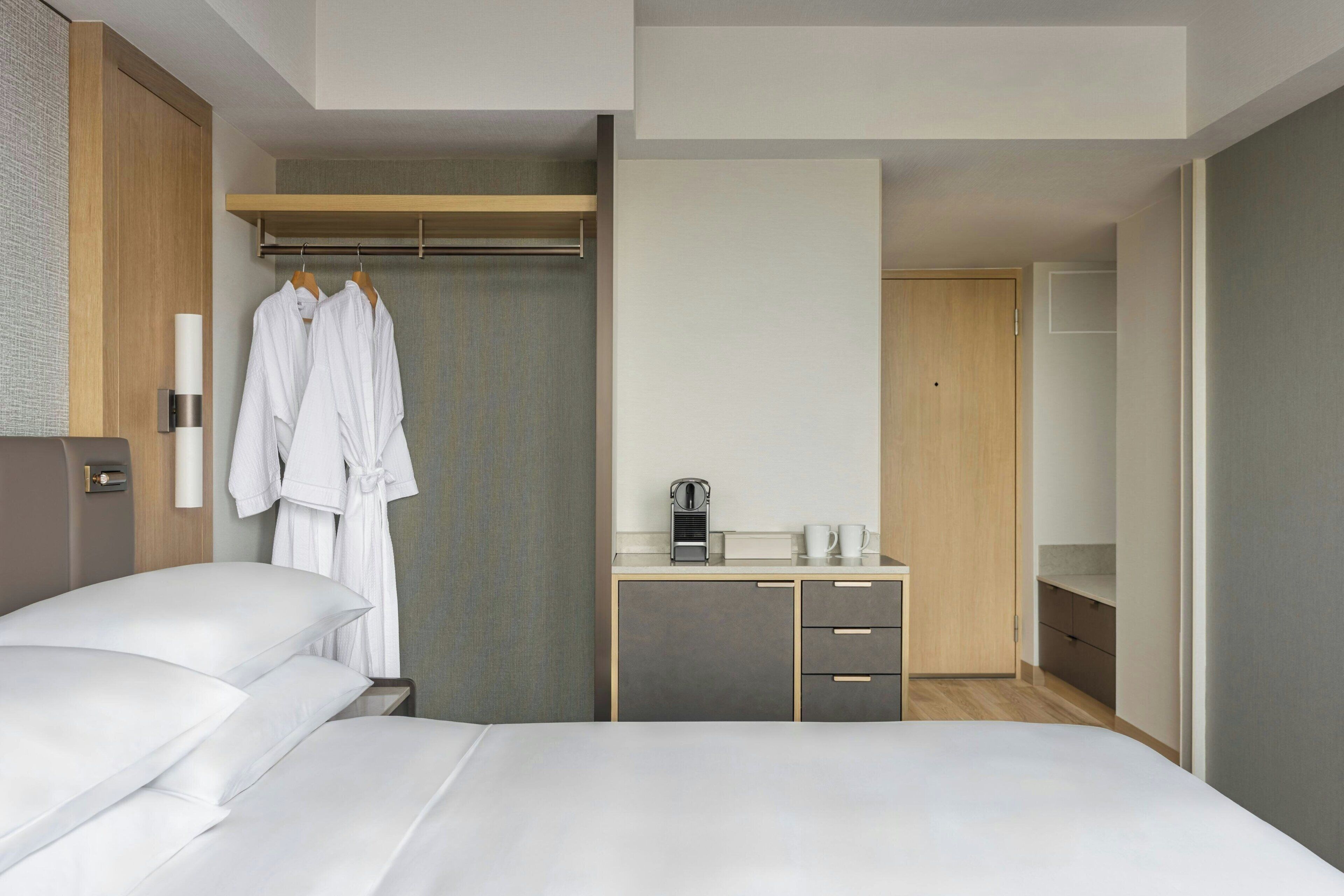 A modern hotel room with a bed, a wardrobe, a desk with a coffee maker, and a mirror.