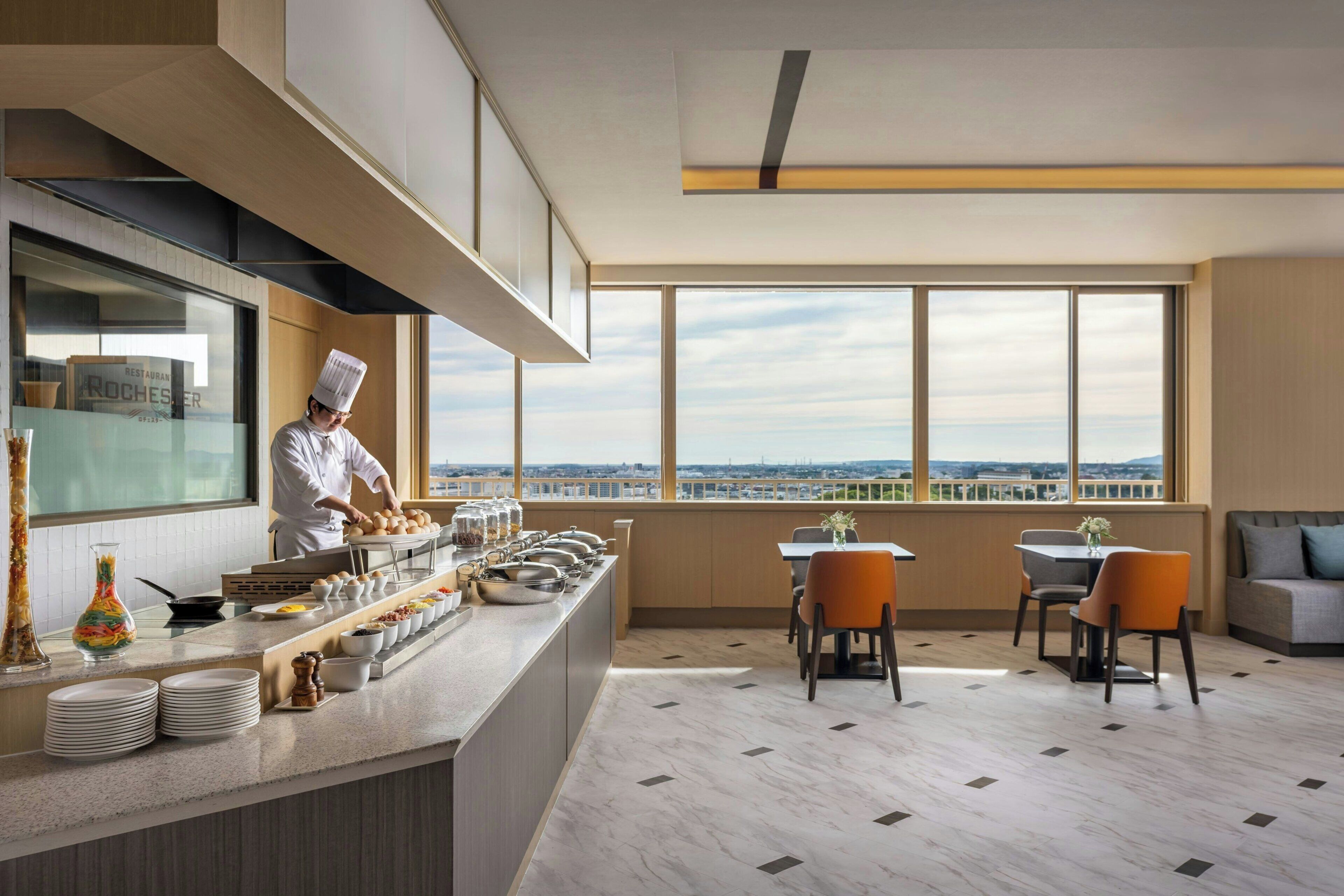 A modern restaurant with a chef preparing food, a buffet with various dishes, and a seating area with a view.