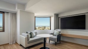 Una habitación de hotel moderna con sofá, sillón, mesa redonda, ventana grande y televisor de pantalla plana.