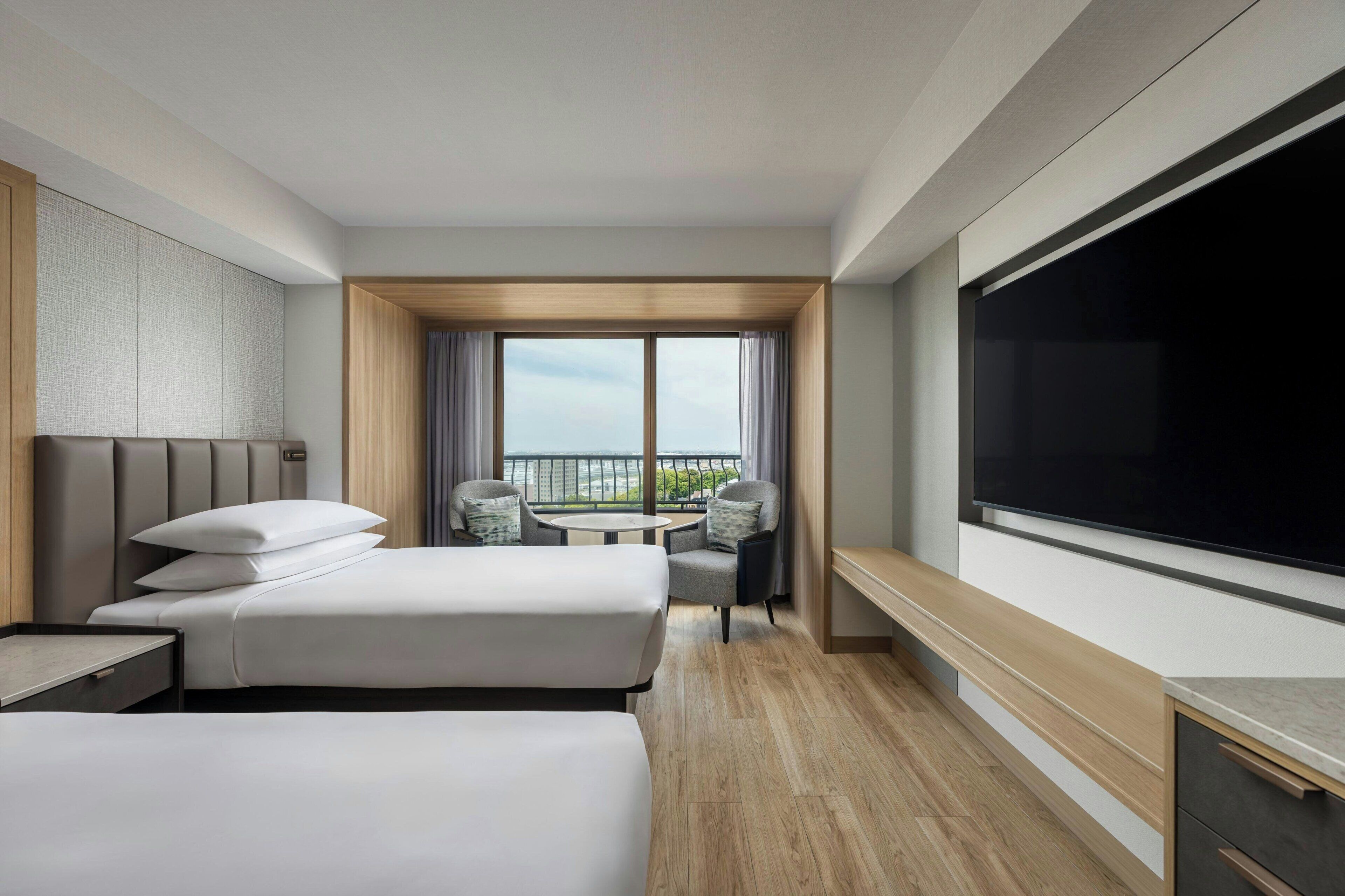 A hotel room with two beds, a TV, a balcony with a view, and a small table.