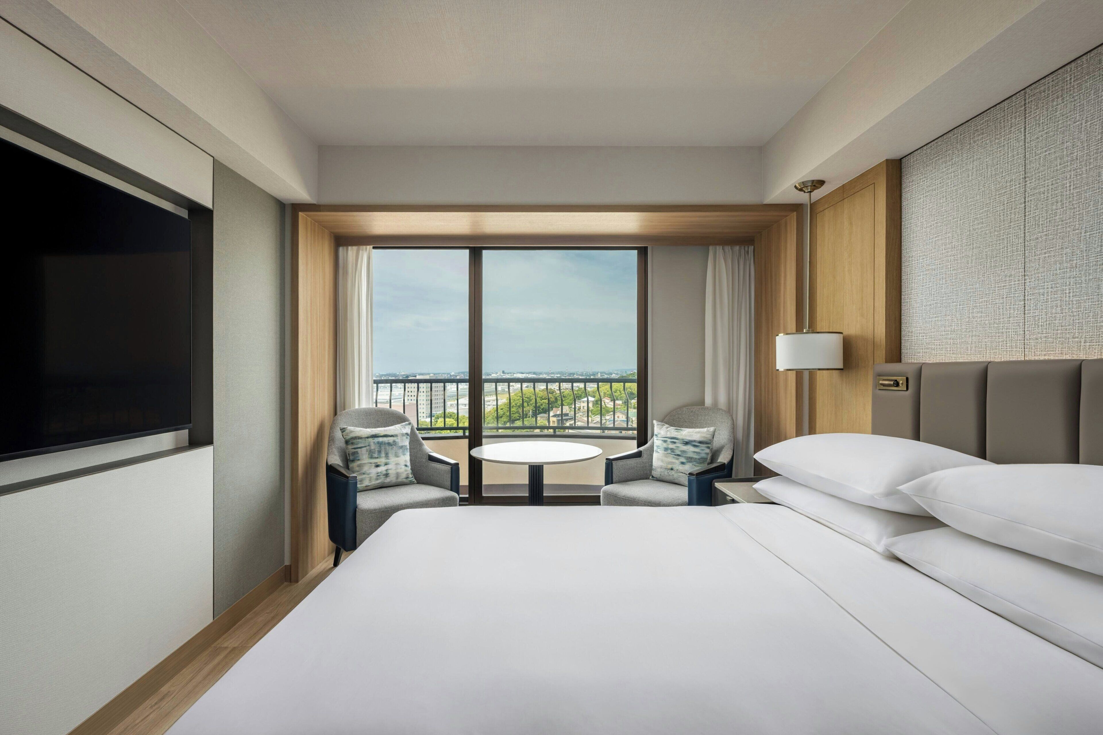 A hotel room with a large bed, a TV, a balcony with a table and chairs, and a city view.