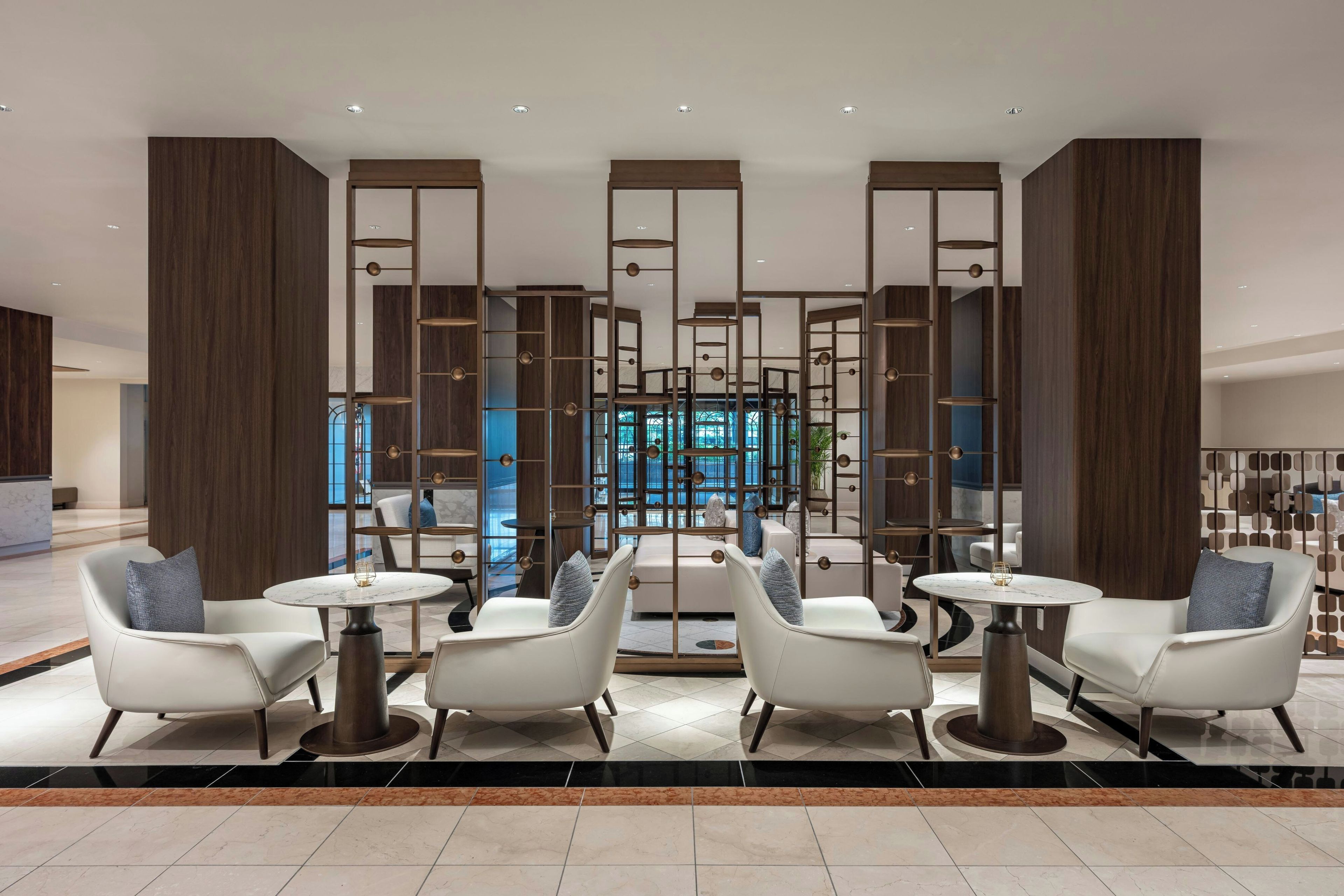 A modern lobby with a seating area featuring white chairs and round tables, a central decorative partition, and a view of the outside through large windows.
