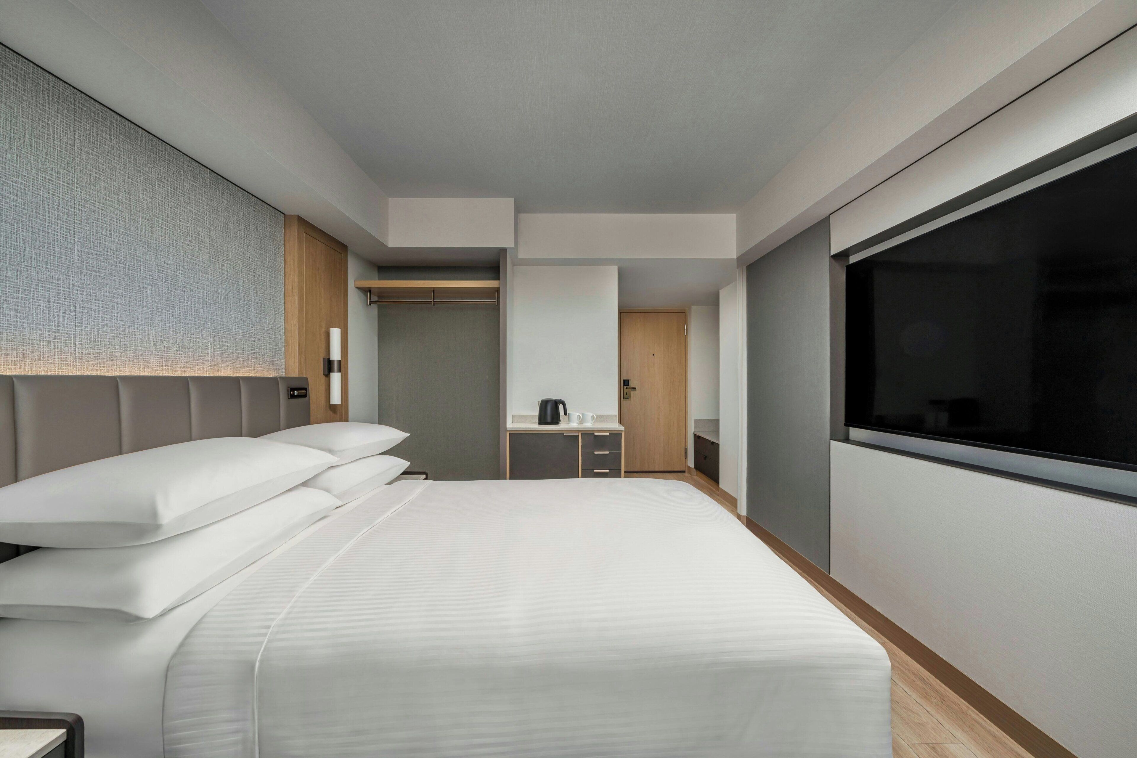 A modern hotel room with a large bed, a small kitchenette, a walk-in closet, and a large window.