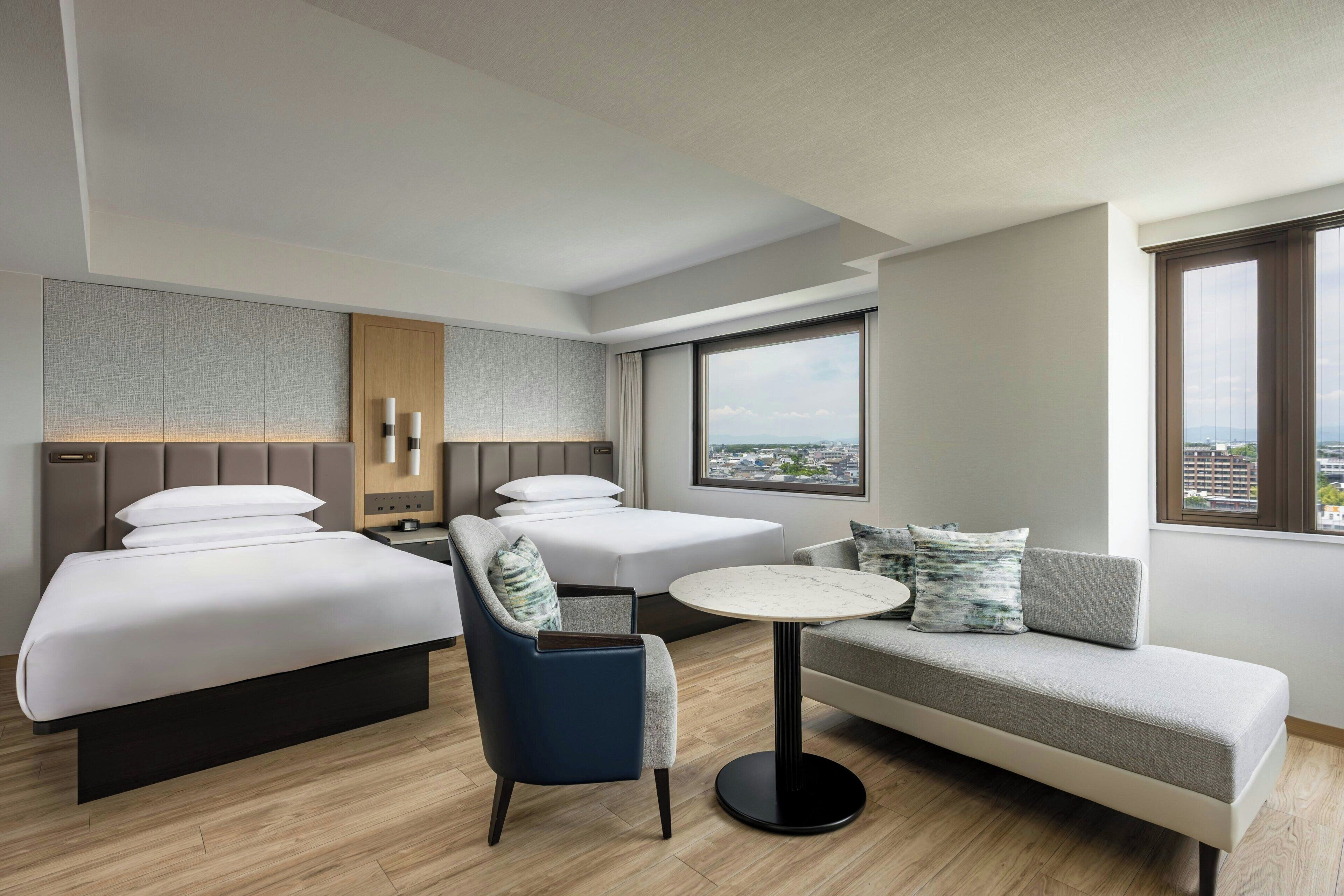 Habitació d'hotel amb dos llits, un sofà, una cadira, una taula petita i una finestra gran que ofereix vista a la ciutat.